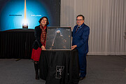 Ein Mann und eine Frau halten ein großes gedrucktes Bild eines Nebels. Im Hintergrund ist rechts ein großer weißer Vorhang zu sehen und links ein hellblaues Schild mit der Aufschrift: Aniversario ESO y Chile.