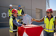 Two people wearing high-visibility jackets and hard hats are standing, holding a silver hexagonal plaque from either side. Behind them, three more people, also wearing construction equipment, look on.