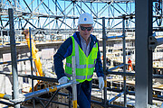 In the midst of a construction site, a person stands at the centre of the frame. They're wearing a high-visibility jacket, a standard white hard hat and sunglasses, and looking directly at the camera. The surroundings include scaffolding, steel beams and various construction equipment.