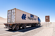 A white and blue lorry, or truck, labelled Hapag-Lloyd is captured on a grey concrete road which stretches off into the distance in the centre right of the photo. A dusty, beige ground sits on either side. The pastel blue daytime sky is in contrast above. To the right of the lorry, a brown roadside sign has an image of a telescope dome with the words Cerro Paranal.
