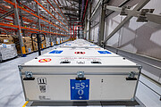 Inside a large warehouse made of a metal structure with white siding, white boxes with the ESO logo — four stars around the letters E, S, O on a blue background — are lined up in a row stretching off into the background. To the left, tall metal and orange shelves sit empty.