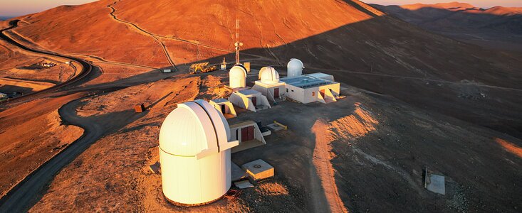 A large, rusty red, sunlit hill dominates the top half of this image. Set below it in the foreground are several white telescope domes and buildings. In the background, atop the hill, is another grey, criss-crossed telescope dome. A large shadow is cast by the hill on the right of the image, where other hills peak into the pastel horizon.