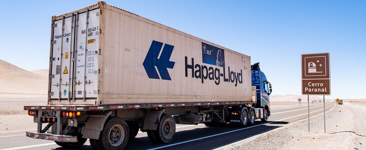 A white and blue lorry, or truck, labelled Hapag-Lloyd is captured on a grey concrete road which stretches off into the distance in the centre right of the photo. A dusty, beige ground sits on either side. The pastel blue daytime sky is in contrast above. To the right of the lorry, a brown roadside sign has an image of a telescope dome with the words Cerro Paranal.