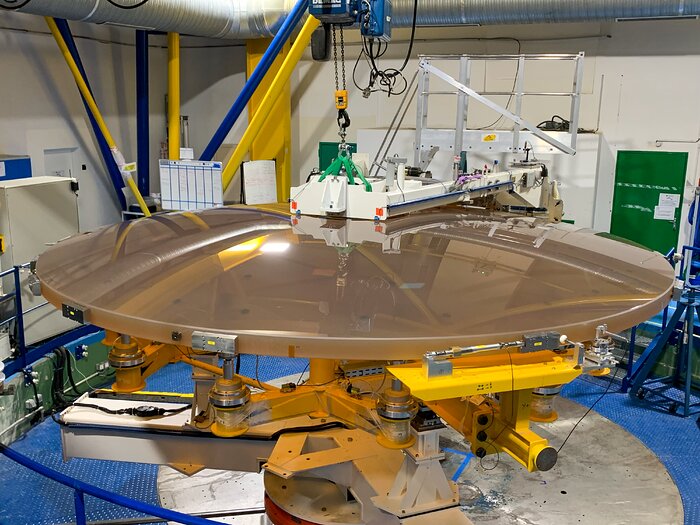 In a workshop setting, a large, shiny, brown-coloured parabolic disc faces towards the floor. The whole dish rests on yellow metal supports. A white measuring device sweeps across the disc’s surface on the far side.