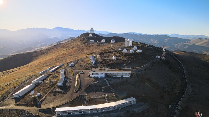 This aerial photograph is taken on a crisp sunny day from high above the La Silla Observatory. Bright white light from the Sun is streaming in from the upper left corner. The light gets fainter away from the Sun, and the sky above is a bright blue in the upper right regions of the image. Beneath the sky mountains stretch off into the distance. On the closest mountain, which is the central focus of the image, long white buildings and smaller circular structures are scattered all around the side of the mountain. The former are the residential and control buildings, located at the bottom of the image. The latter are the many telescopes found here, located up the mountain slightly from the buildings.
