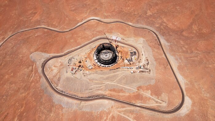 This drone image looks down on orange-brown ground, with a dark road spiralling into the centre of the image. The criss-cross circular steel structure of the dome sits at the centre of the spiral, while construction equipment is dotted around its circumference.