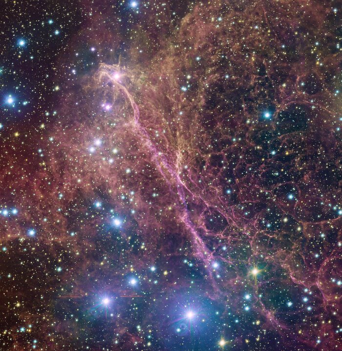 At the centre of the image, there is a pink network of filaments, elongated from upper-left to lower-right. In the upper part of these filaments, in the opposite direction, from upper-right to lower-left, filamentary orange clouds fill the space. Nearby, dark red filaments and clouds fill the space. Some clouds have well-defined filamentary shape, others are more blurred and cloudy-shaped. Spread all over the picture, bright yellow, blue and reddish stars populate the image. The dark background is almost completely hidden by all these features.