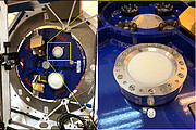 This image is in two parts, split vertically down the centre: on the left there is a Band 2 receiver, and on the right a closeup of the optic lens. On the left is a blue and circular metal panel, surrounded by a grey circular structure. On the blue panel, a number of devices are fitted. Surrounding one of them is a superimposed yellow box, with two lines expanding to the right-hand image. The right hand image is a closeup of what is in the yellow box on the left: it is the warm optics lens of the Band 2 receiver. Circular in shape, the lens is white in the centre and is surrounded by a metal frame, fitted in place with bolts around its circumference.