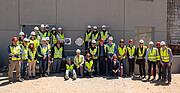 Around 30 people wearing high-visibility jackets and hard hats, some sitting and some standing, are gathered in front of a large grey concrete wall. The metallic time capsule and plaque are fitted into the wall in the middle of the group.