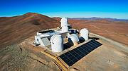 Several white telescope domes sit on a flattened brown hilltop. Angled slightly away from the camera, three sit in the front row, one in the middle connected to a white building, and one in the back.  A set of black solar panels rest in front of the domes. In the background, darker brown hills roll off into the distance under a vivid blue sky.