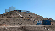 In the foreground of this image, a cube container is branded with ‘DLR’. One person is climbing a ladder to reach the top of the container, whilst another watches nearby. There is another plain blue shipping container close by. In the background of the image, white buildings sit high atop a hill on the desert landscape.