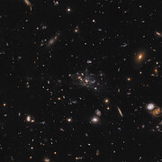This image shows several galaxies distributed over a black background. In the centre of the image is a larger concentration of galaxies. The galaxies have colours that range from blue, orange-red, yellow and white.