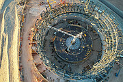 This aerial photograph, taken soaring above the ground using a drone, shows the construction site of the Extremely Large Telescope, ELT. The site is basking in golden sunlight that is creeping in from this side of the image. The circular structure of the telescope is almost directly below the camera and is made up of steel beams, criss-crossing each other. At the centre of the circle a white crane stands tall, pointing off to the left side of the image. One gets a sense of how big the ELT structure is when comparing it to a white car, parked just left of centre at the top of the image — it is tiny compared to this colossal structure. Along the left hand side a brown dirt road winds around the building.