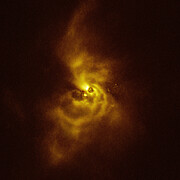In this image a ghostly cloud lurks centrally in the frame on a dark background. The yellow and brown, wispy cloud extends up and down from a central bright region to the edges of the frame, making an elongated spiral shape that gets dimmer and less defined as it gets further from the centre.