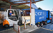 A person sitting in a grey and orange transporter loads two large crates onto a blue truck. The truck sports white text reading “SCHOTT goes ELT”, with the ESO logo — four stars surrounding the letters E, S, O, all in white — next to it. The text on the crates reads “SCHOTT” in blue letters.