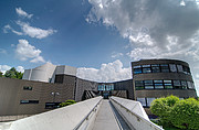 Image showing the ESO headquarters on a bright sunny day from the concrete bridge leading towards the main reception. The building features large curved walls made of brown corrugated metal and concrete, with lots of windows.
