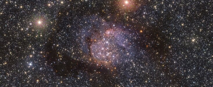 Dieses Bild ist mit unzähligen Sternen übersät, die als leuchtende weiße Punkte von unterschiedlicher Größe und Helligkeit vor dem schwarzen Hintergrund des Weltraums erscheinen. In der Mitte des Bildes befinden sich weitere Sterne, die in eine violette Wolke eingetaucht sind. Um sie herum befindet sich der Serpens-Nebel, der auf diesem Bild als schwaches, dunkles, orangefarbenes Glühen erscheint. Durch die Wolke hindurch sind jedoch noch einige Sterne sichtbar.