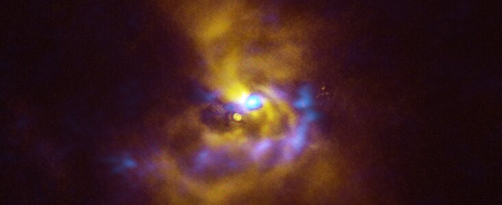 The background of this image is dark, but in its centre lurks a swirling ghostly figure, which extends towards the edge of the picture. At the very centre there is a small bright region and erupting out of it there is a poorly defined, fuzzy edged cloud and blobs of material in yellow and blue, respectively. The yellow cloud extends far out in the image, making an elongated spiral shape that gets dimmer and less defined as it reaches the top and bottom of the frame. Meanwhile, the blue blobs only extend downwards from the centre and to a fraction of the distance of the yellow spiral cloud. The blobs twist away from the central bright region, forming a tight U-shape lying on its right side.