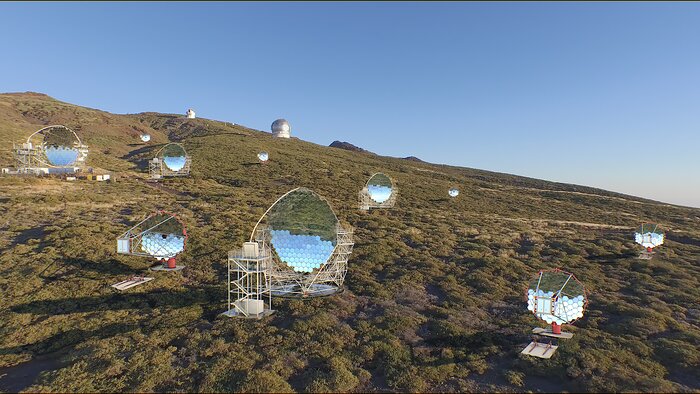 An artist’s impression of the CTAO-North telescope array under the sunny blue sky of La Palma, Spain. Several reflective parabolic dishes of varying sizes are dotted along the green hillside, which slopes down from left to right. The dishes themselves are made up of numerous hexagonal segments. In the background, further up the slope, a couple of white telescope domes that are not a part of CTAO can be seen peeking over the brow of the hill.