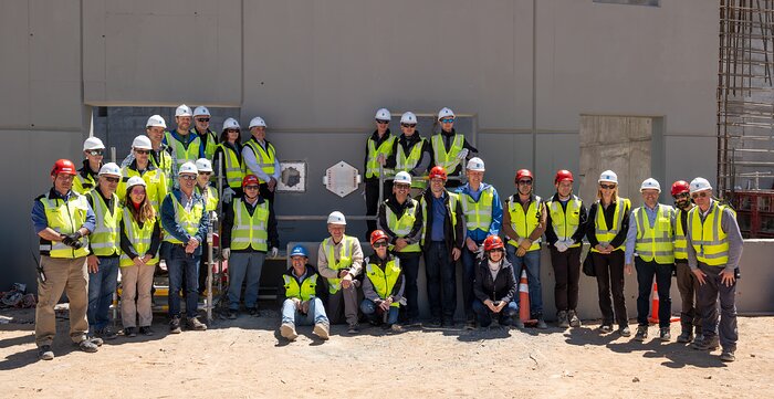 Around 30 people wearing high-visibility jackets and hard hats, some sitting and some standing, are gathered in front of a large grey concrete wall. The metallic time capsule and plaque are fitted into the wall in the middle of the group.