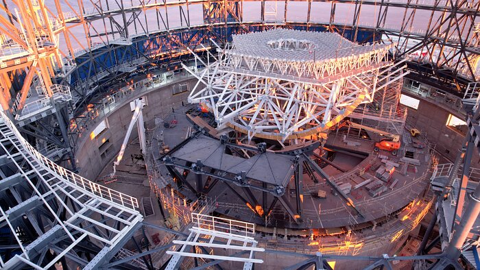 This image is taken inside a metallic dome structure. In the centre of the dome, a white lattice honeycomb-like structure is under construction in a hexagonal shape, surrounded by construction equipment, cranes, and vehicles.