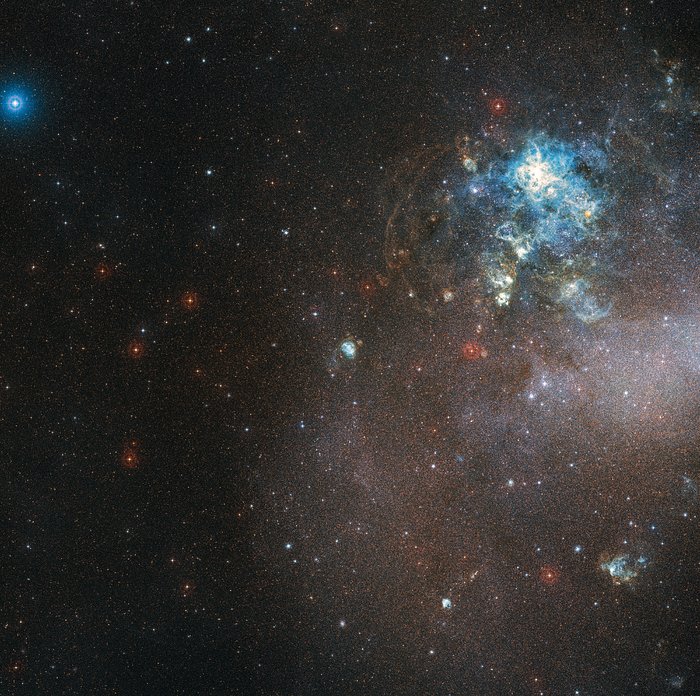 The image shows a dark area of the night sky, peppered with white, red and blue dots of starlight. The right hand side of the image is brighter and milkier. Towards the upper right lies a mesmerising, wispy region of clouds, coloured most strikingly by a region of electric blue at the centre of the clouds.