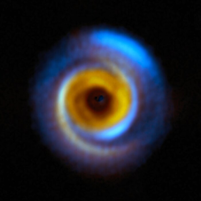 Ein kreisförmiges, dunstiges und farbenfrohes Objekt befindet sich auf einem schwarzen Hintergrund. Zu seinen äußeren Rändern hin hat es spiralförmige Arme in Blau und Orange. Nach innen hin hat es einen helleren orangefarbenen Ring und einen dunkelroten inneren Kreis. In der Mitte befindet sich ein schwarzer Fleck.