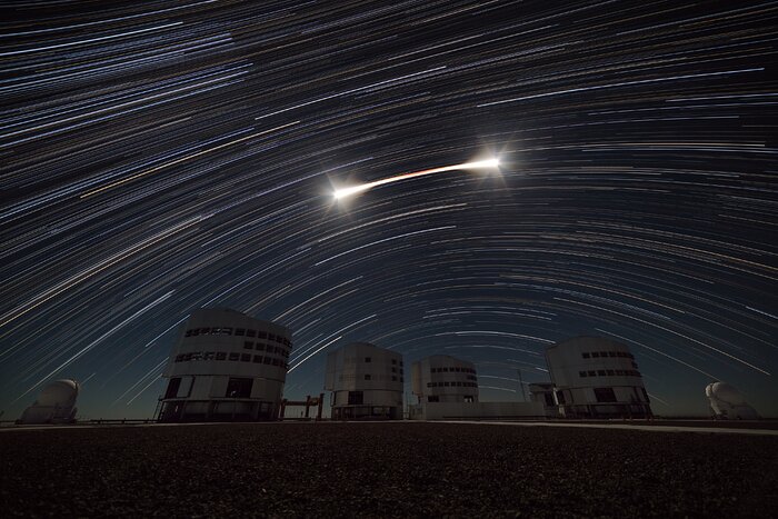 The image shows the light from the moon as a long bright streak in the middle of the picture. The streak has two bright points at both ends, and a reddish hue in the middle. Around the streak are elongated star trails. The trails fill the majority of the image. At the lower part of the image are the four VLT telescopes.