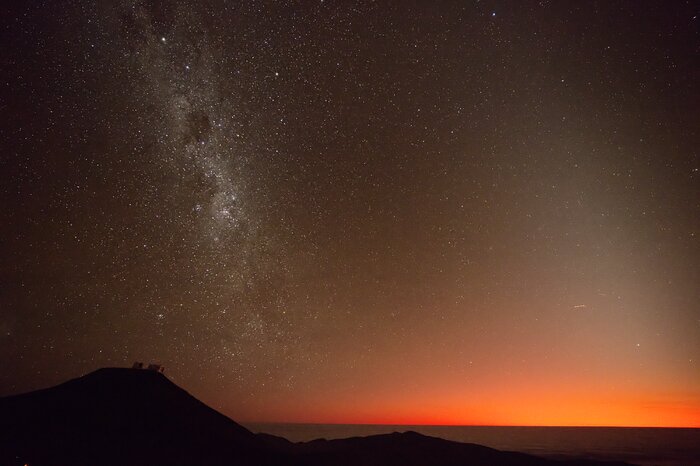 This picture was taken at ESO’s Paranal Observatory at sunset. The silhouette of Chilean mountains occupies the lower part of the picture, with a higher peak on the left and one lower on the right. Above, the red-orange twilight sky overlooks the picture. The sky is darker on the left, where the Milky Way appears.