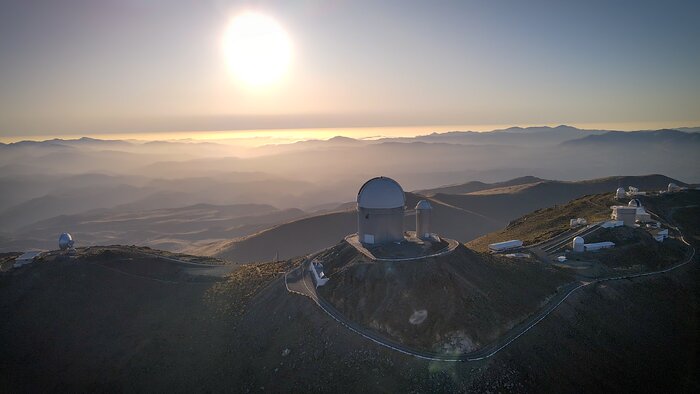Two different sceneries are layered in this image. In the background, the mountains, almost hidden by a whitish veil, create a fairy tale atmosphere. In the foreground, the ridge of the mountains hosts several telescopes on its smooth peaks. On the leftmost peak there is only one telescope. In the central and highest one, there is one big telescope, also alone. And finally, the rightmost peaks are crowded with smaller telescopes and facility buildings. The background and the foreground are immersed in the warm light of the Sun, which fires in the centre of the whitish sky.
