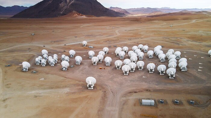 A light caramel plateau spans across almost all of the image. The monotony of the plateau is broken by smooth hills and here and there dirt roads appear whitish. Half of the image is occupied by ALMA’s white antennas, closely packed together on the plateau. The photo is taken from the back of the antennas, with both larger 12-metre and smaller 7-metre dishes present. Farther away, on the left, there is a massive dark brown mountain, whose peak is not part of the frame. Beyond this giant are other brown and caramel mountains. The sky occupies only a very small portion of the image, and it is cloudy and whitish.