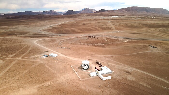 A coffee and caramel coloured landscape spans across almost all the picture. The flatness is broken by smooth hills and, here and there, by occasional whitish dirt roads. In the lower central region of the picture there is the large white APEX telescope and some ancillary buildings, all surrounded by white fencing. The two white buildings on the left of APEX are quite close to the telescope. Further away, in the upper right part, ALMA’s antennas appear as white dots close to each other. Further still, the brown mountains extend throughout all the width of the image. Above them, the cloudy sky occupies only a small band of frame.