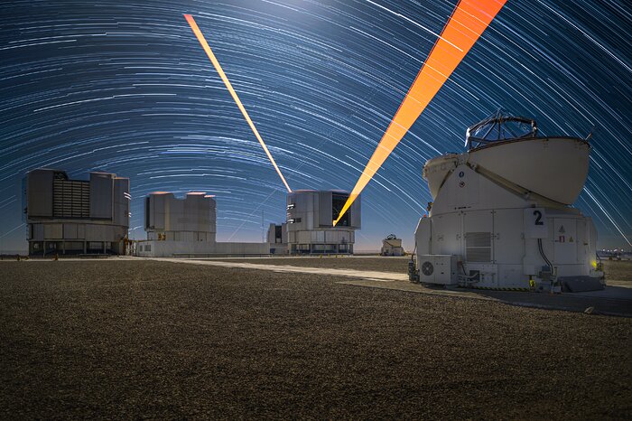 A swirling, dynamic, long-exposure image of Paranal Observatory at night. The backdrop of the night sky is a deep blue, with long arcs of white light bending across it horizontally. The circular pattern of stars is broken by two beams of orange light erupting out of a cylindrical telescope house. There are two other similar cylindrical domes to its left, and a smaller one further behind. In the foreground of the image on the right-hand side is the spherical white structure of an Auxiliary Telescope, with its shell open so that the telescope inside can observe the cosmos.