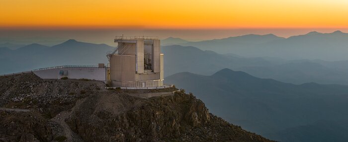 This image, which looks more like a painting than a photograph, was captured at sunset. Across the top, a band of orange sky runs from left to right. The focal point of the image is the New Technology Telescope dome, a large cylindrical grey structure. The shutter doors of the dome are slightly parted. It’s perched high on a rocky mountain top, surveying more shadowy mountains that fill the background below the sky.