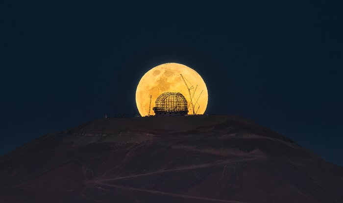 The yellow disc of the full moon stands out against the dark backdrop of the night sky. The moon has nearly risen above the flat mountain top at the centre of this image, but is just cut off from being a perfect circle at the bottom. Like shadow puppets on a screen, the criss-crossed steel structure of the ELT dome is silhouetted at the centre of the moon, accompanied by towering cranes.