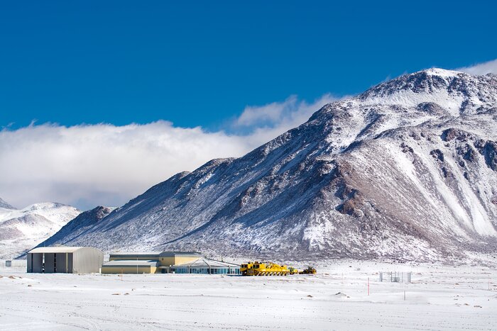 A photograph of a snowy landscape. The top third is taken up by a bright blue sky, the middle third shows mountains in front of white clouds, and in the bottom third some low buildings and a bright yellow transporter stand on the flat, snowy terrain.