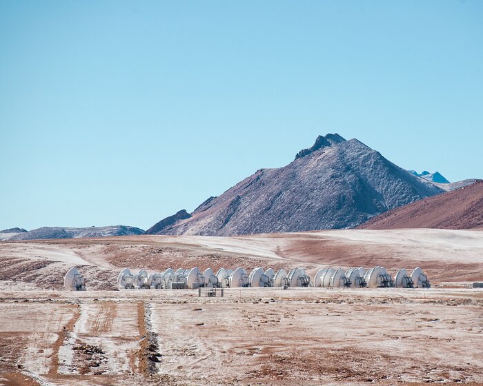 The photo is of the landscape of the Chajnantor Plateau, in the Chilean desert. Mountain ranges are in the background, and the sky is blue. In the distant center-point of the photo, many ALMA antennas —large white “dishes” — are packed tightly in a cluster.