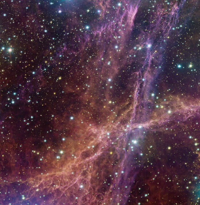 The image shows the remnants of a supernova explosion, which appears as a wispy structure of pink and orange clouds. While there are clouds, which look like thin filaments, all over the image, one major pink column can be seen going from the top of the image to the bottom, meanwhile an orange coloured column of clouds stretches from left to right in the bottom half of the image. Throughout the image there are also many stars, shining with white, orange and blue light. Some of these stars are much brighter and larger than others.
