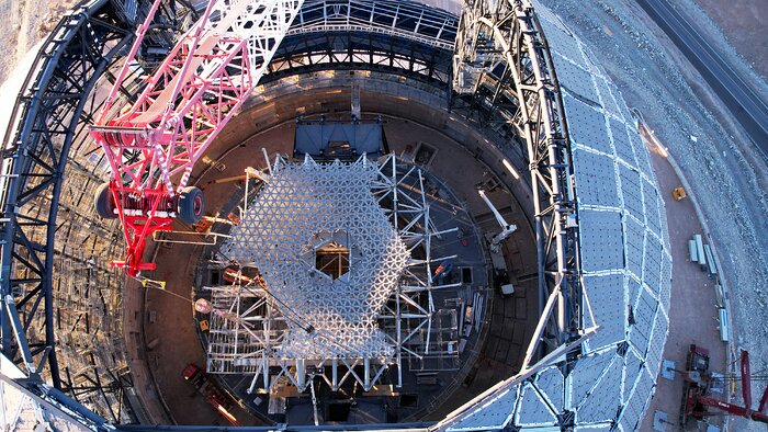 The image is a bird’s-eye view of a partly-constructed circular metallic structure, without a top. A red and white crane is peaking into frame at the top of the image. Inside the dome, a white honeycomb lattice structure is being constructed in a hexagonal shape, with a central hole.
